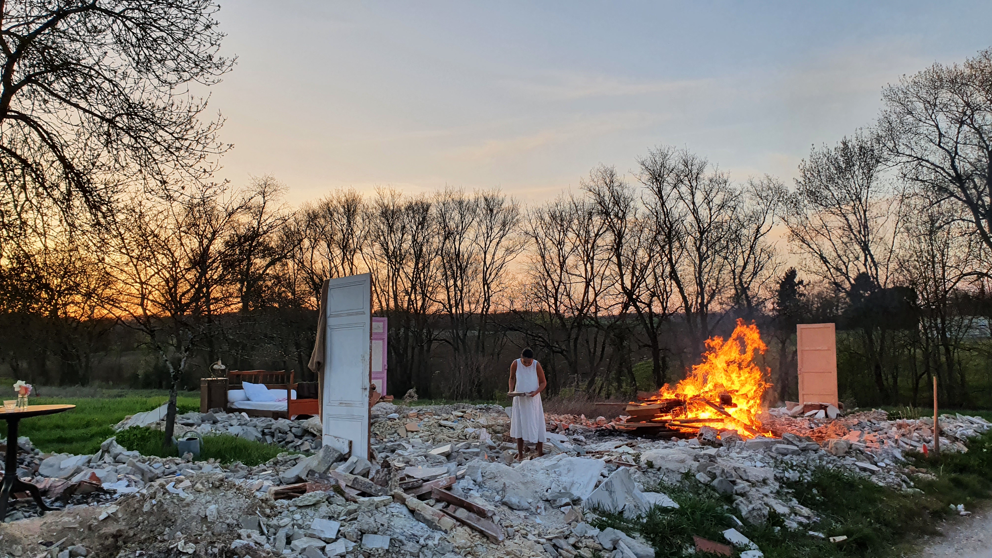 A man stands amidst a pile of rubble and burning debris in an outdoor setting at sunset, with various pieces of furniture ...
