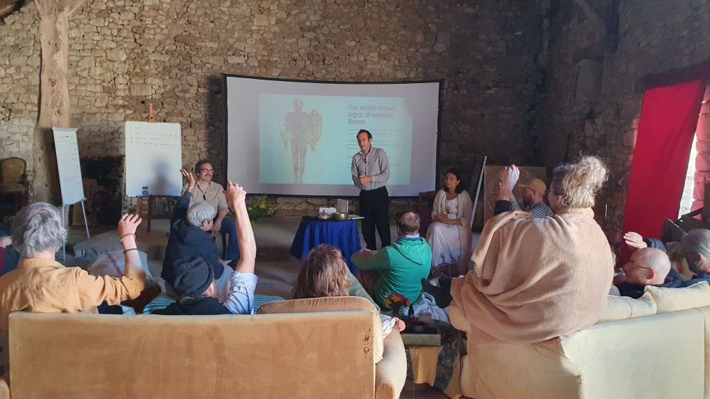 A man gives a presentation to an audience. His slide includes the caption: "Our world shows signs of serious illness". The...