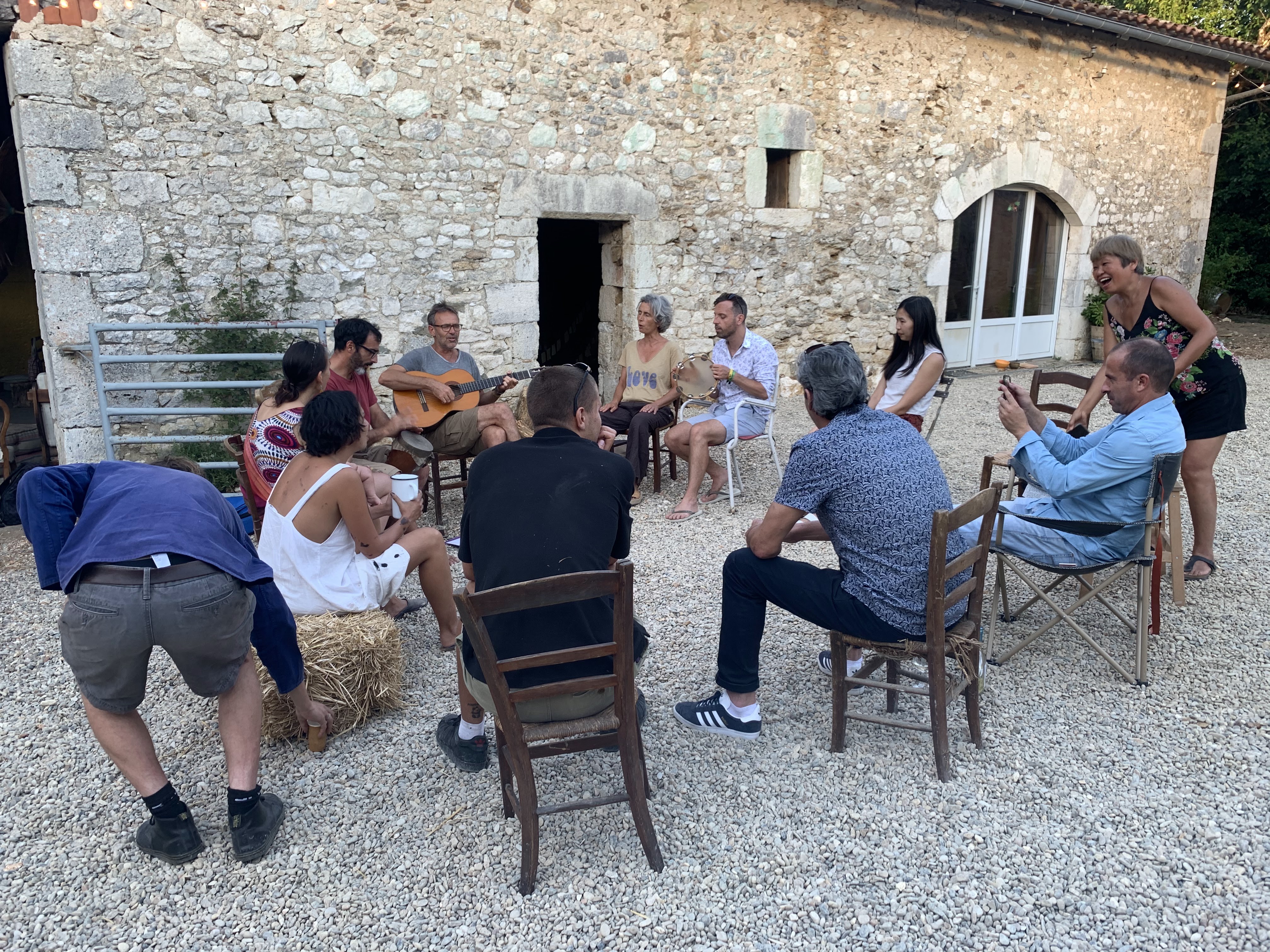 A group of people sitting outside an old stone building enjoying one another's company. Roughly half-women and half-men ar...